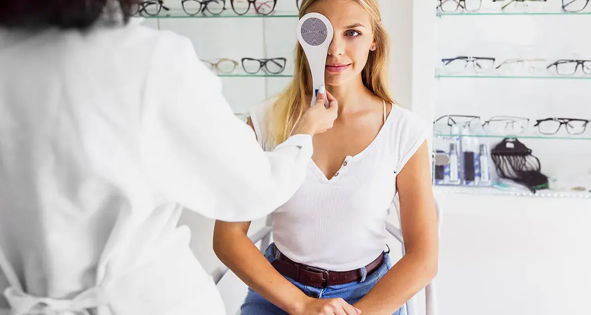 Młoda kobieta przechodzi badanie wzroku. Optometrysta zasłania jej jedno oko przyrządem do cover testu.