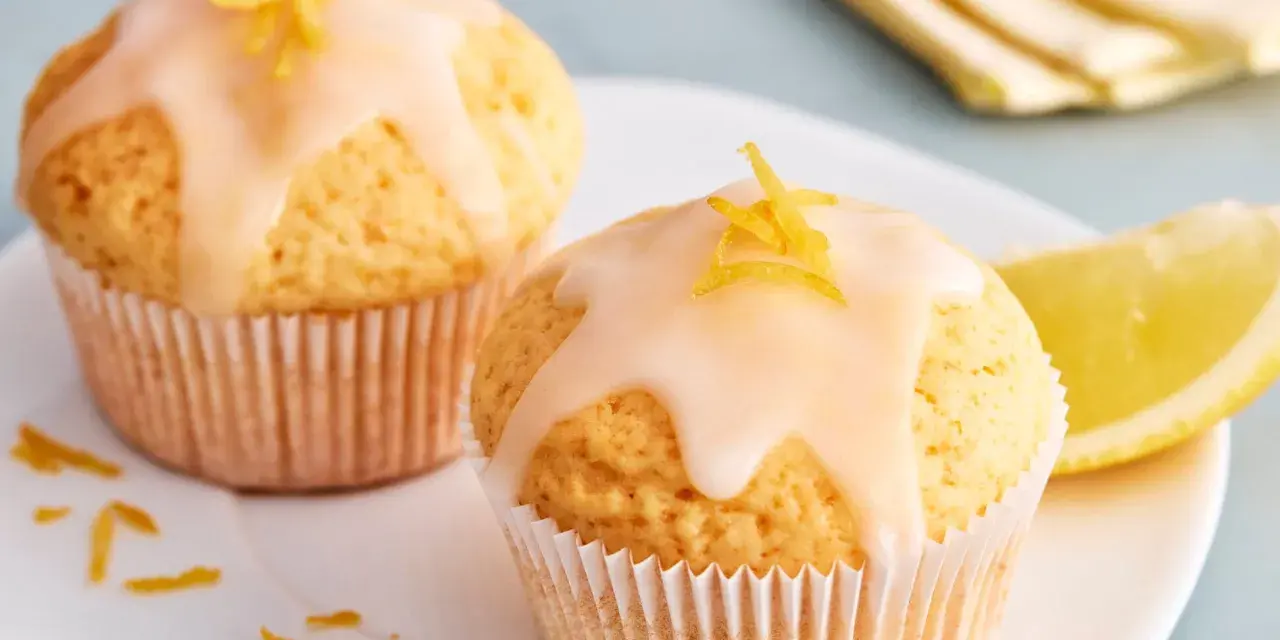 Zwei einfache Zitronenmuffins mit Zuckerguss und Zitronenschale garniert, daneben ein Zitronenschnitz.