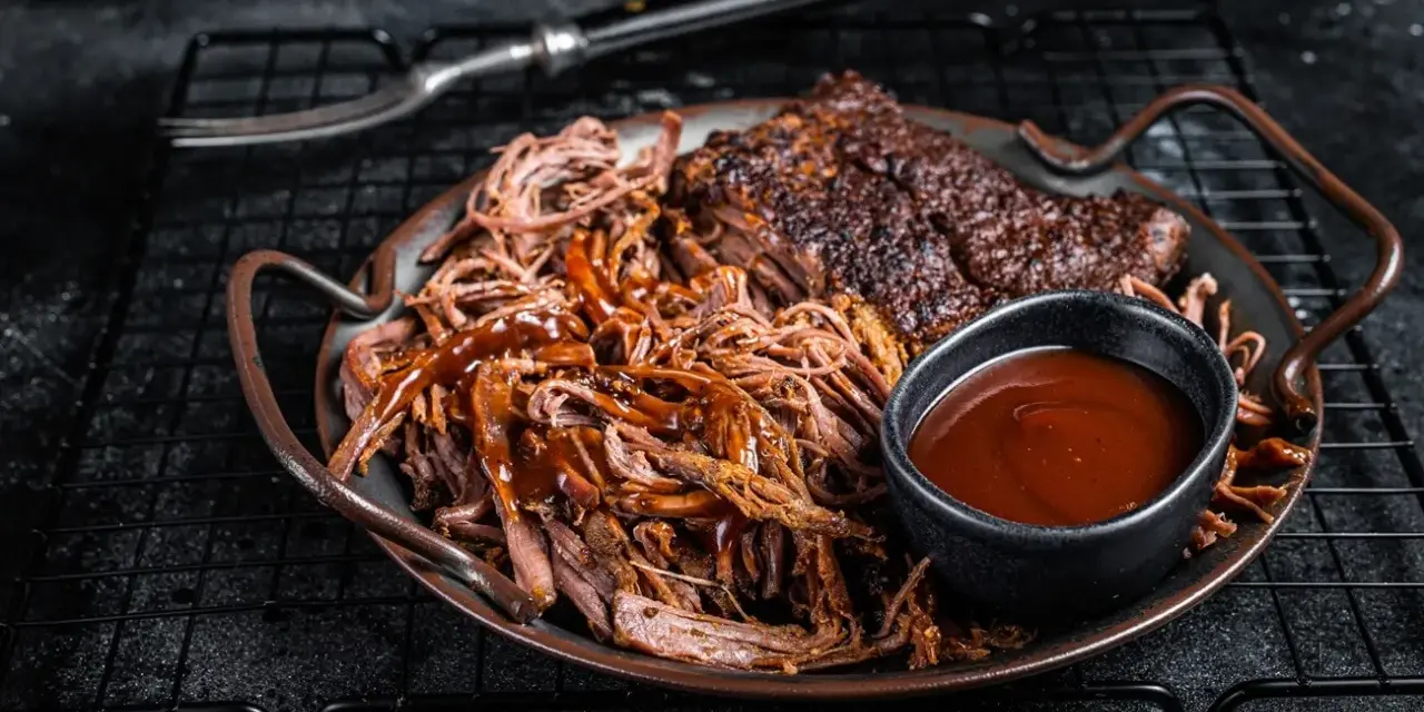 Saftiges pulled pork mit BBQ-Sauce und Beilagen auf einem Teller. Ein Gabel liegt daneben.