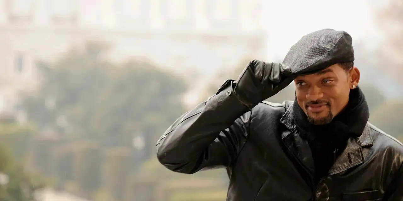 Will Smith, con un berretto uomo, giacca di pelle e sciarpa, saluta con un sorriso.