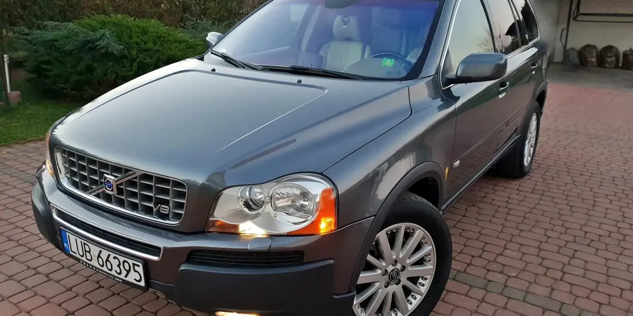 Szary Volvo XC90 z silnikiem V8, zaparkowany na podjeździe z kostki brukowej, otoczony zielenią.