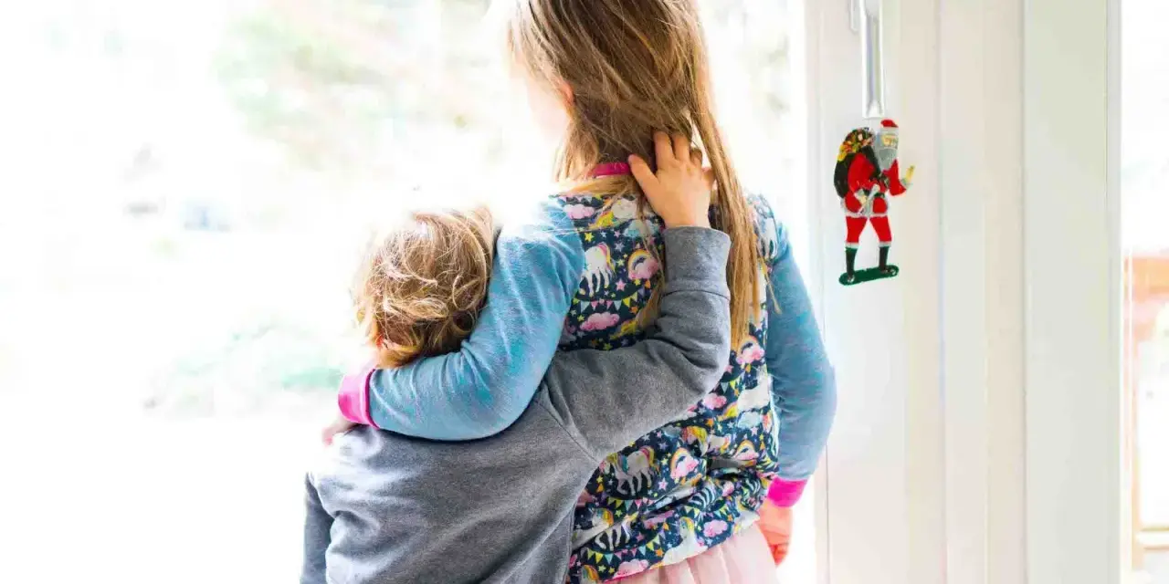 Zwei Kinder umarmen sich vor einem Fenster. Das jüngere Kind, das Angst hat, wird vom älteren Kind getröstet. Ein Weihnachtsmann-Anhänger hängt an der Tür.