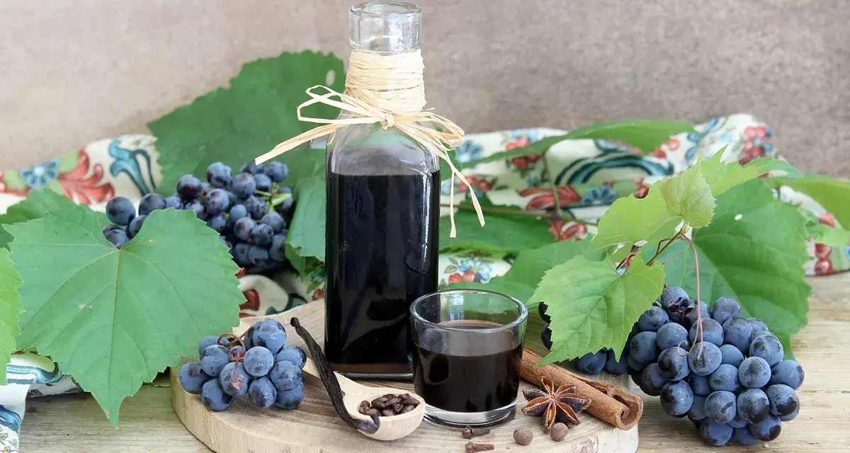 Mosto cotto ricetta: sciroppo scuro in bottiglia e bicchierino, con uva, spezie e cucchiaio di semi.