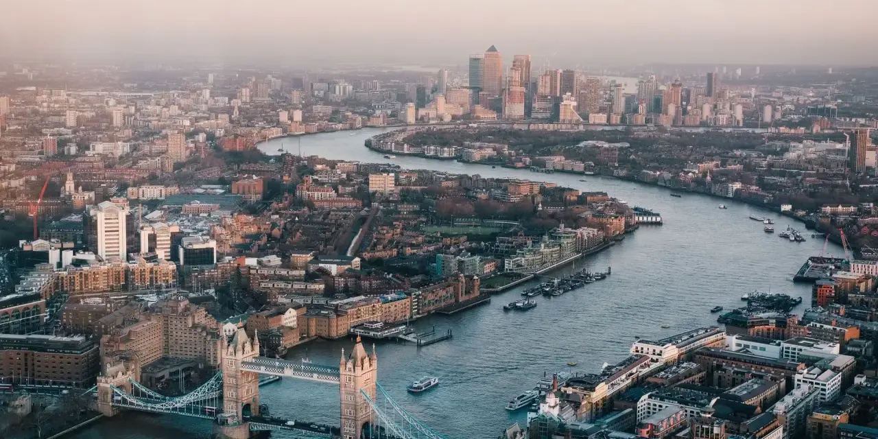 Stunning drone photography and videography captures London's iconic Tower Bridge and the winding Thames River, with the city skyline stretching into the hazy distance.