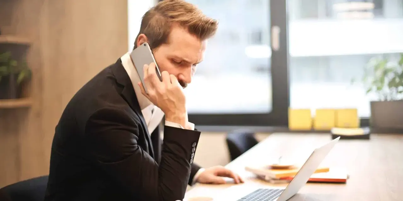 Mężczyzna w garniturze prowadzi ważną rozmowę przez telefon, siedząc przy biurku z laptopem i filiżanką kawy.
