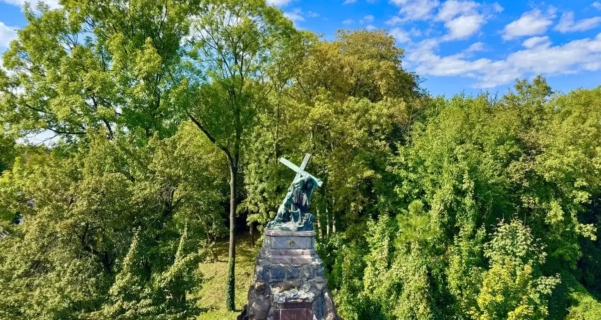 Pomnik na Jasnej Górze w Częstochowie, otoczony bujną zielenią drzew, pod błękitnym niebem z chmurami.