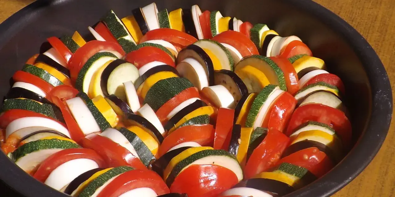 Un plat de légumes coupés en rondelles, prêt pour une recette de ratatouille digne du film. Tomates, courgettes, aubergines et oignons s'alternent.
