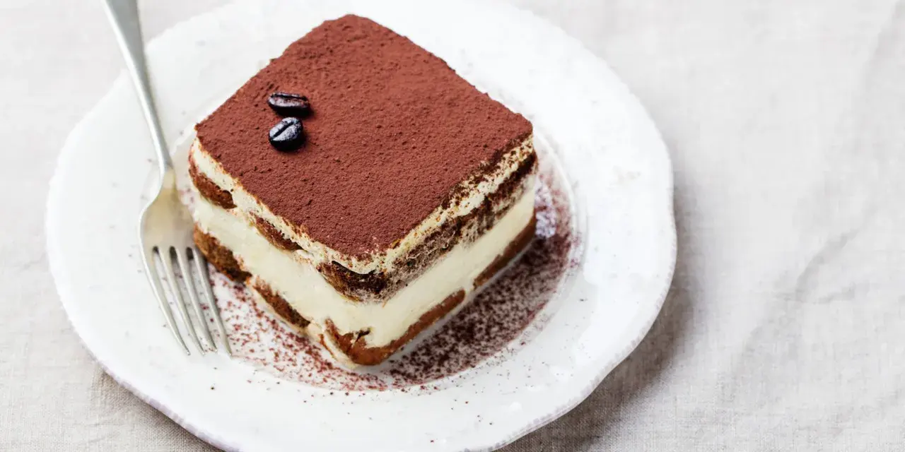 Una fetta di tiramisù, preparata con 500 g di mascarpone, guarnita con cacao e chicchi di caffè, su un piatto bianco con forchetta.