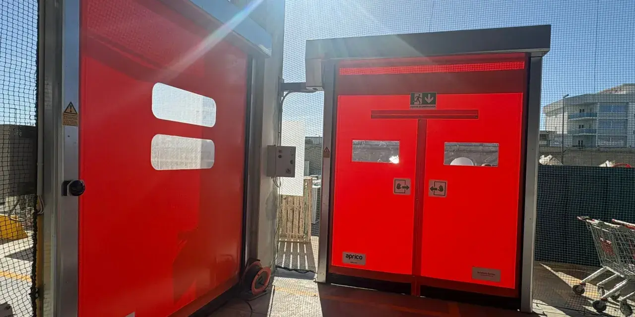 Due porte rosse automatiche, una aperta, mostrano come alzare una porta dal pavimento. Luce solare intensa.
