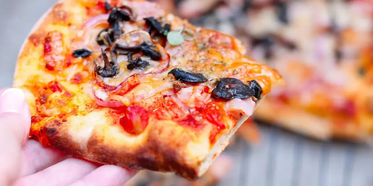 Kawałek pysznej pizzy funghi z pieczarkami, cebulą i pomidorami, trzymany w dłoni.