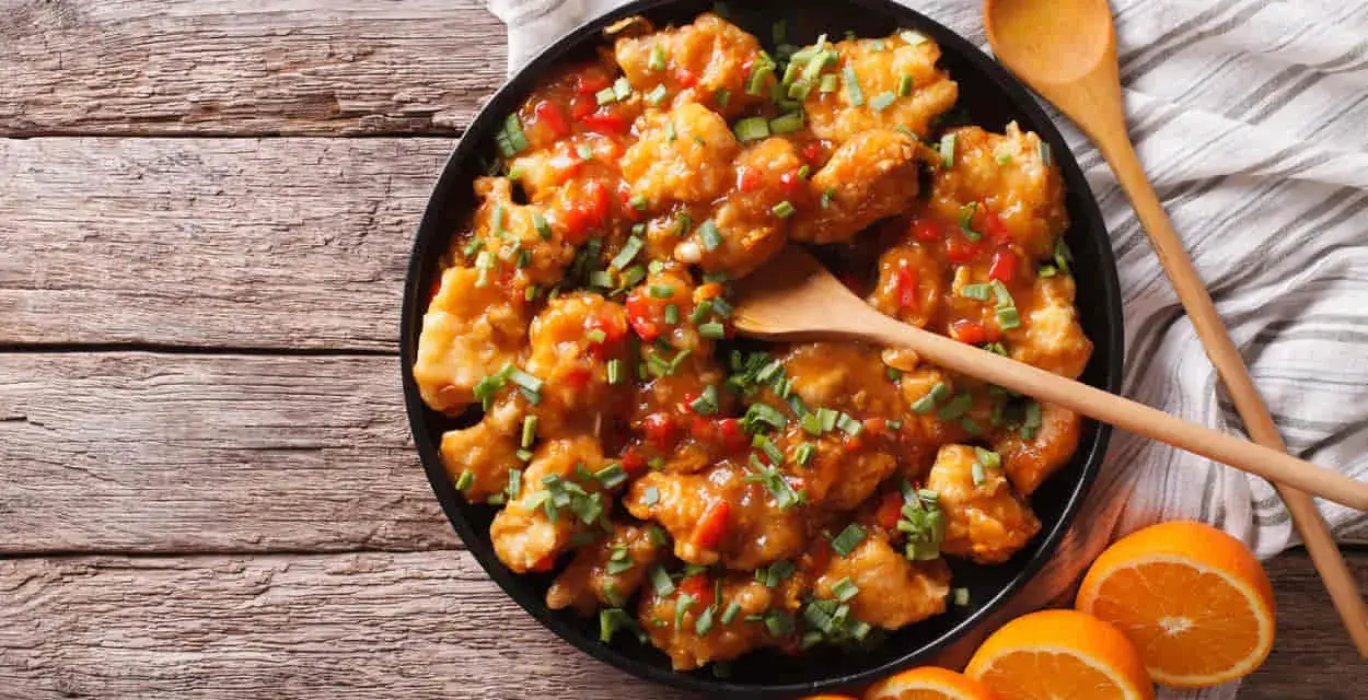 Sabroso pollo a la naranja, cubierto con salsa brillante y cebollín picado, servido con rodajas de naranja fresca.