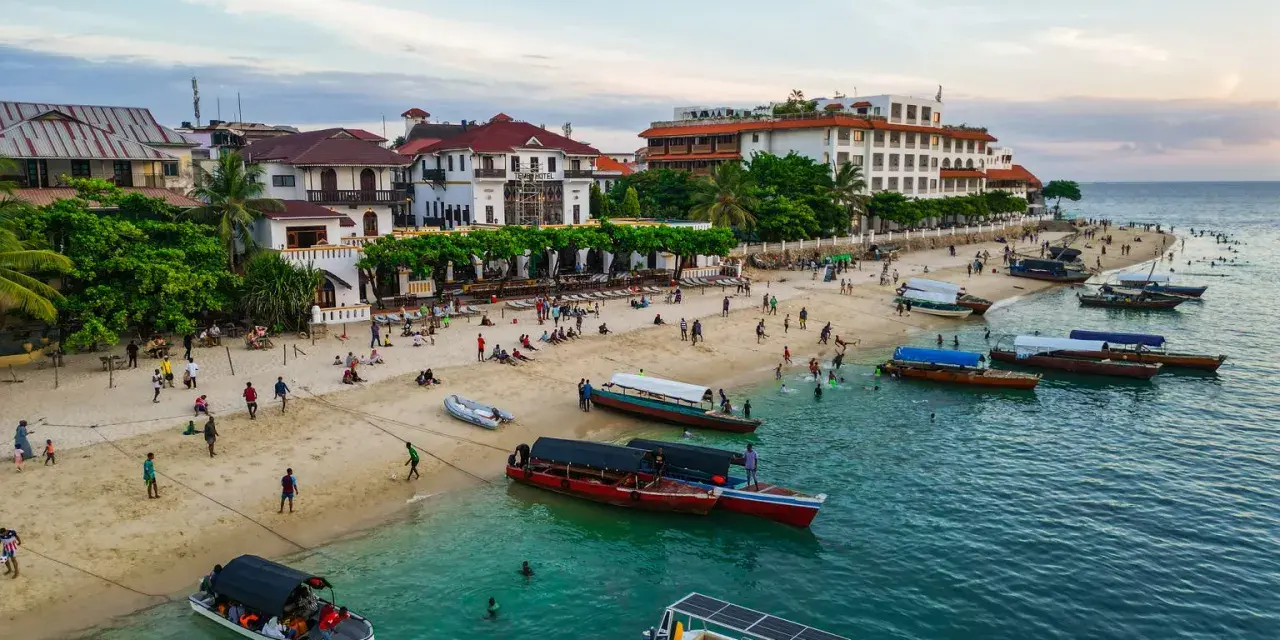 Tanzański szyling na plaży w Stone Town, Zanzibarze. Ludzie odpoczywają, bawią się i pływają, a łodzie czekają na turystów.