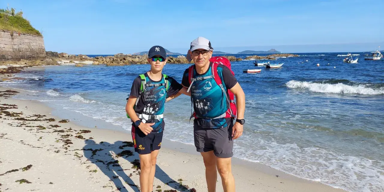 Dos excursionistas, padre e hijo, disfrutan de la playa en la etapa Baiona Vigo. El mar azul y el cielo despejado acompañan su aventura.