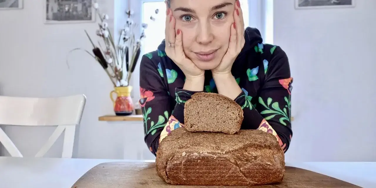 Kobieta z dłońmi podpartymi pod brodą patrzy na pyszny chleb wieloziarnisty. Jeden bochenek leży na drugim, na drewnianej desce.