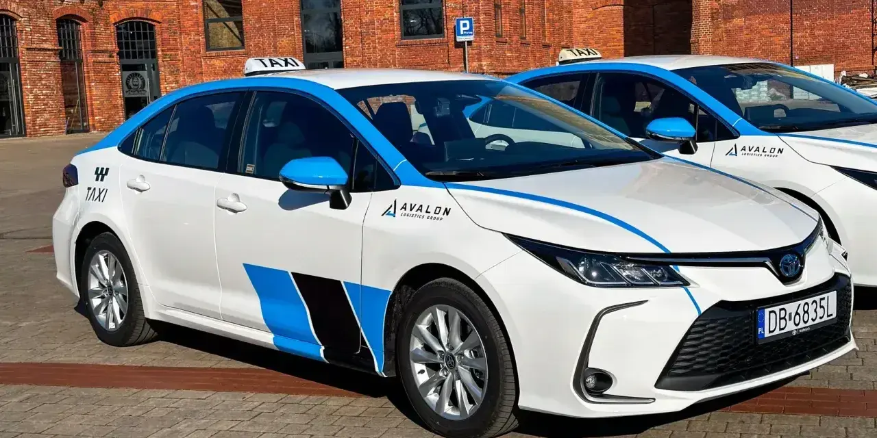 Białe taksówki z niebieskimi pasami i logo Avalon, spełniające uber wymagania samochodu, stoją przed ceglanym budynkiem.