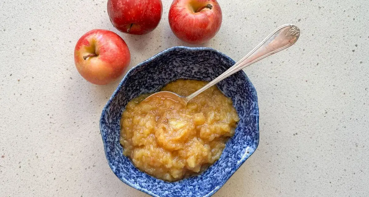 Compota de manzana casera en un cuenco azul, lista para disfrutar. Tres manzanas rojas frescas acompañan este delicioso postre.