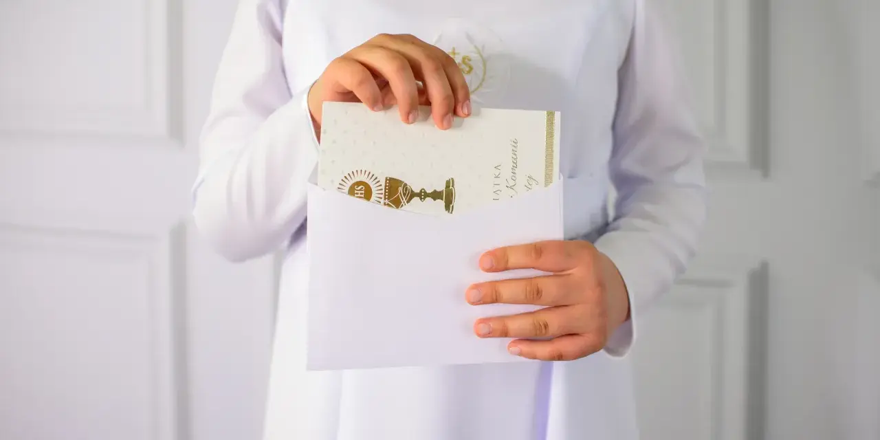 Dziecko w bieli wkłada kartkę z kielichem do koperty. To piękne krótkie życzenia na komunię, pełne symboliki.