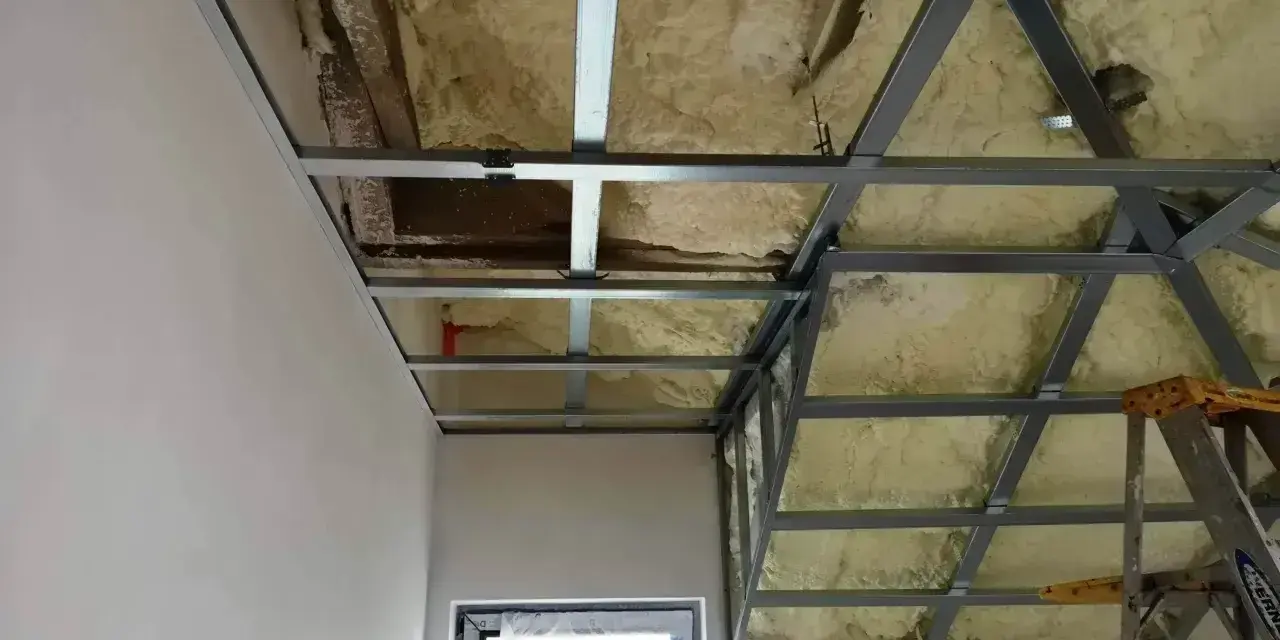 Konstrukcja podwieszanego sufitu z metalowymi profilami i pianką izolacyjną. Widać fragmenty ścian i okna.