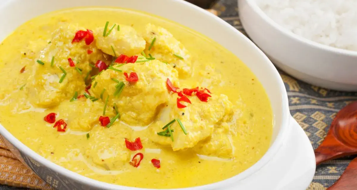 Delizioso pollo al curry con latte di cocco, guarnito con peperoncino rosso e erba cipollina. Accanto, una ciotola di riso bianco.