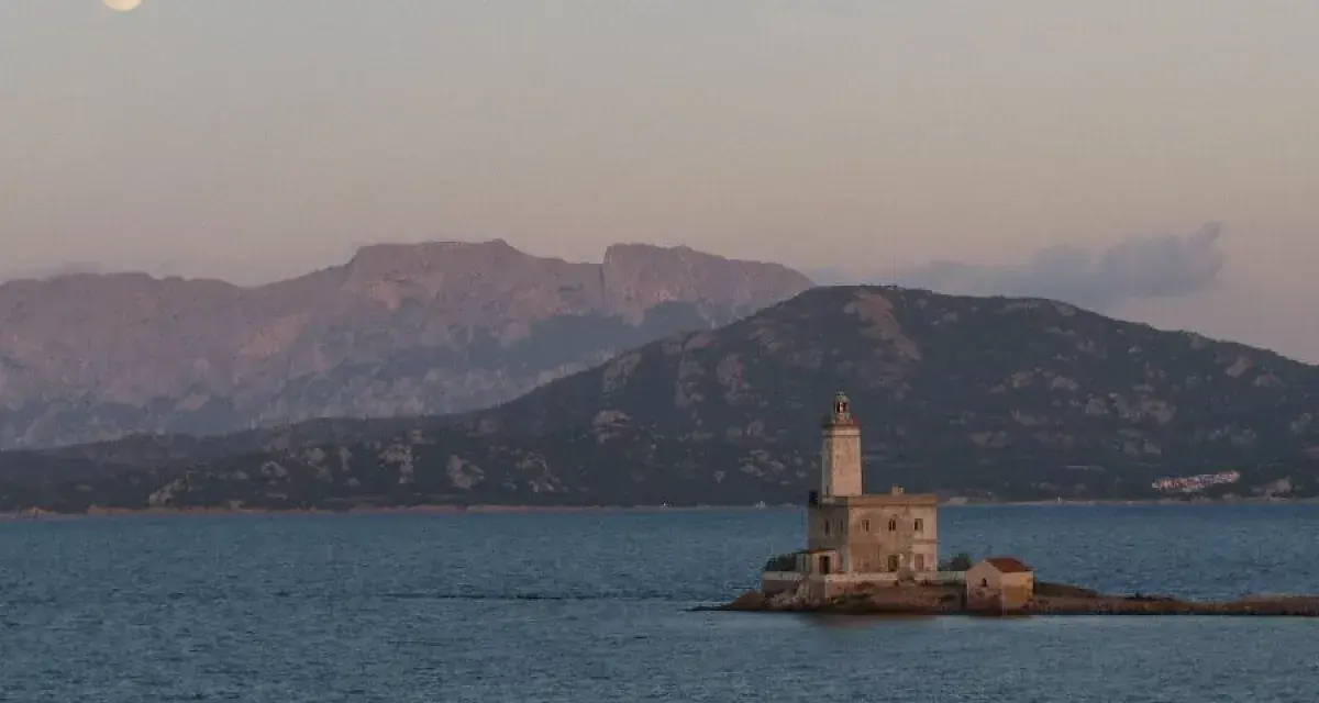 Faro solitario su un'isola sarda, con montagne sullo sfondo e luna piena. Perfetto per itinerari moto Sardegna 7 giorni.