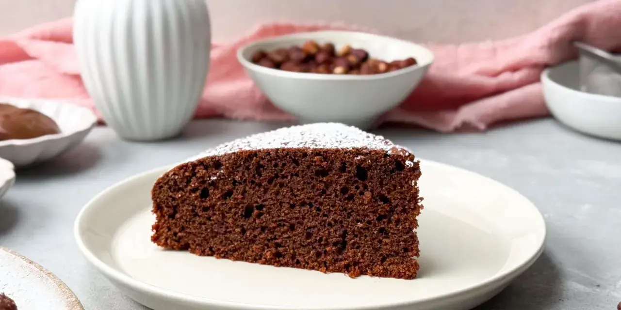 Ein saftiges Stück **Nutella Kuchen** mit Puderzucker bestäubt, daneben Nüsse und eine Vase.