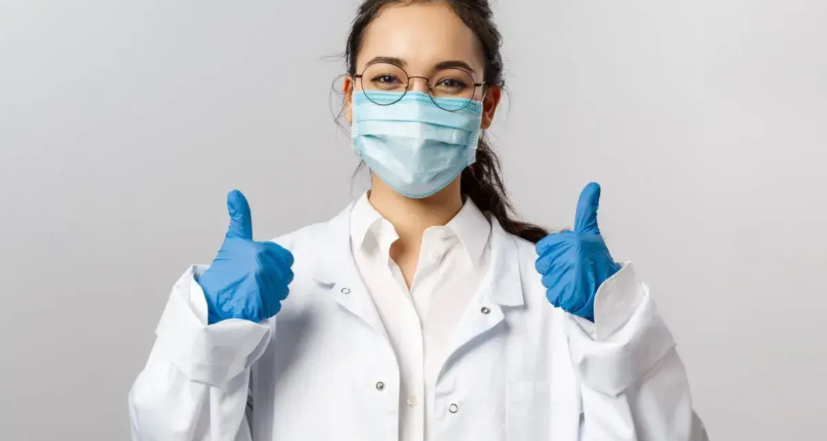 Uśmiechnięta pielęgniarka epidemiologiczna w masce i rękawiczkach pokazuje kciuki w górę.