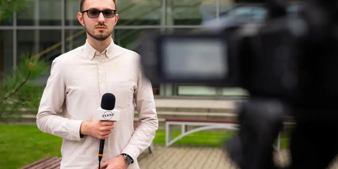 Młody dziennikarz w okularach trzyma mikrofon z logo "FLESZ" przed kamerą.