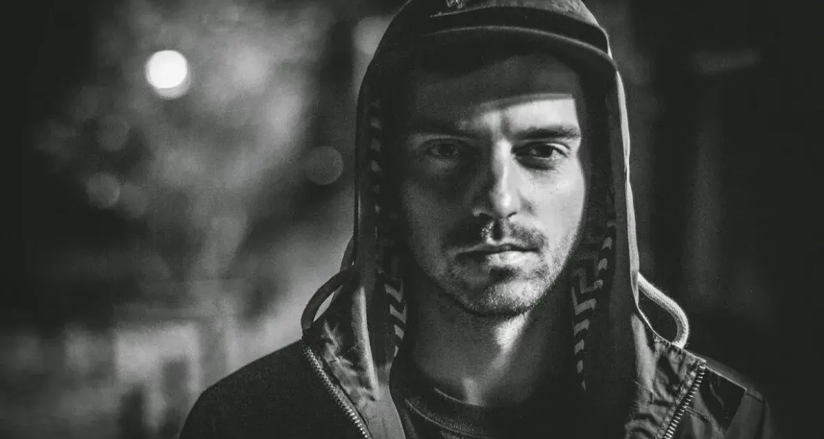 Mroczny portret mężczyzny w kapturze, z zacienioną twarzą i spojrzeniem, które zdaje się mówić o brudnych sercach i trudnych doświadczeniach.