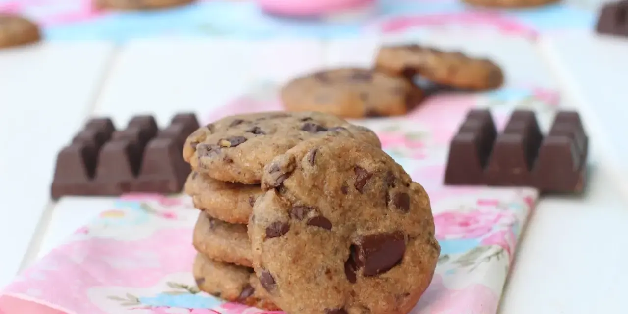 Una pila de galletas de chocolate caseras, con trozos de chocolate visibles, junto a barras de chocolate y un vaso de leche rosa.