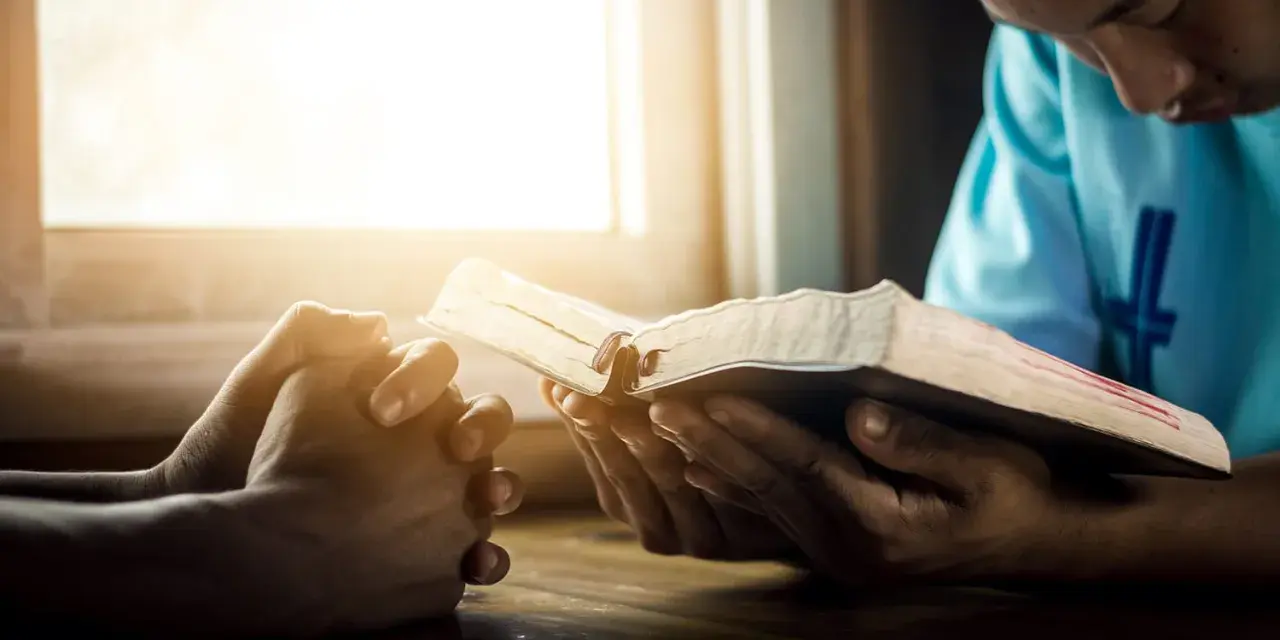 Hands clasped in prayer beside an open Bible, bathed in warm light. A moment of reflection, perhaps contemplating prayer names.