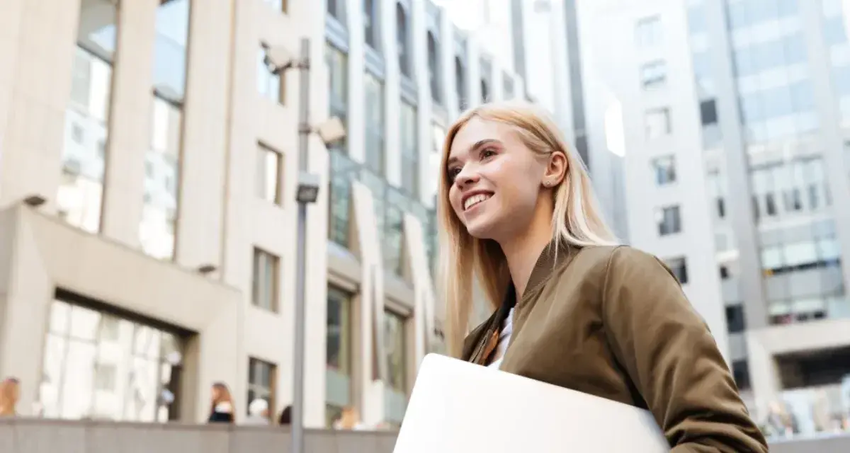 Giovane donna sorridente con laptop, cammina in una città moderna. Lavorare in Svizzera conviene, un futuro brillante ti aspetta.