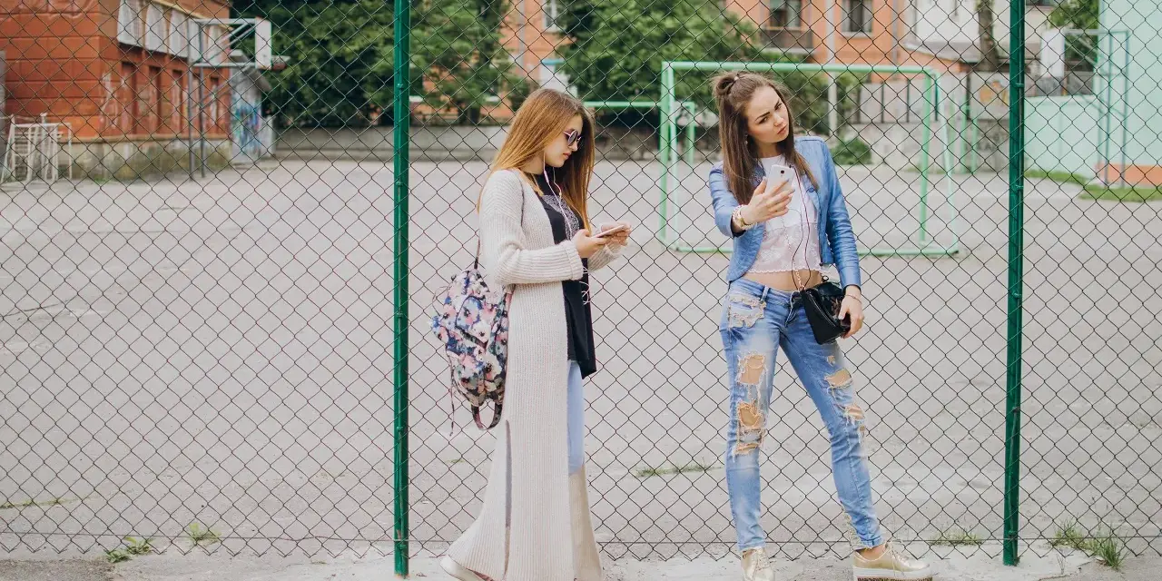 Dwie dziewczyny w modnych stylizacjach, idealnych na outfit do szkoły, stoją przy płocie. Jedna robi sobie selfie, druga przegląda telefon.
