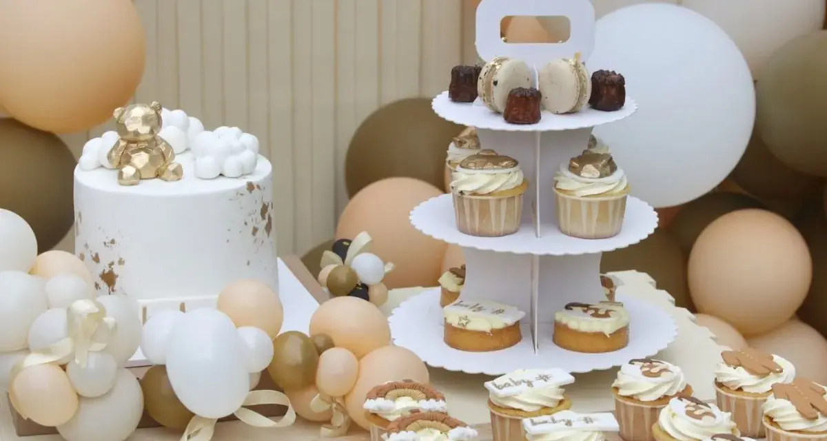 Biały tort z złotym misiem, babeczki i makaroniki na paterze, otoczone balonami w pastelowych kolorach.