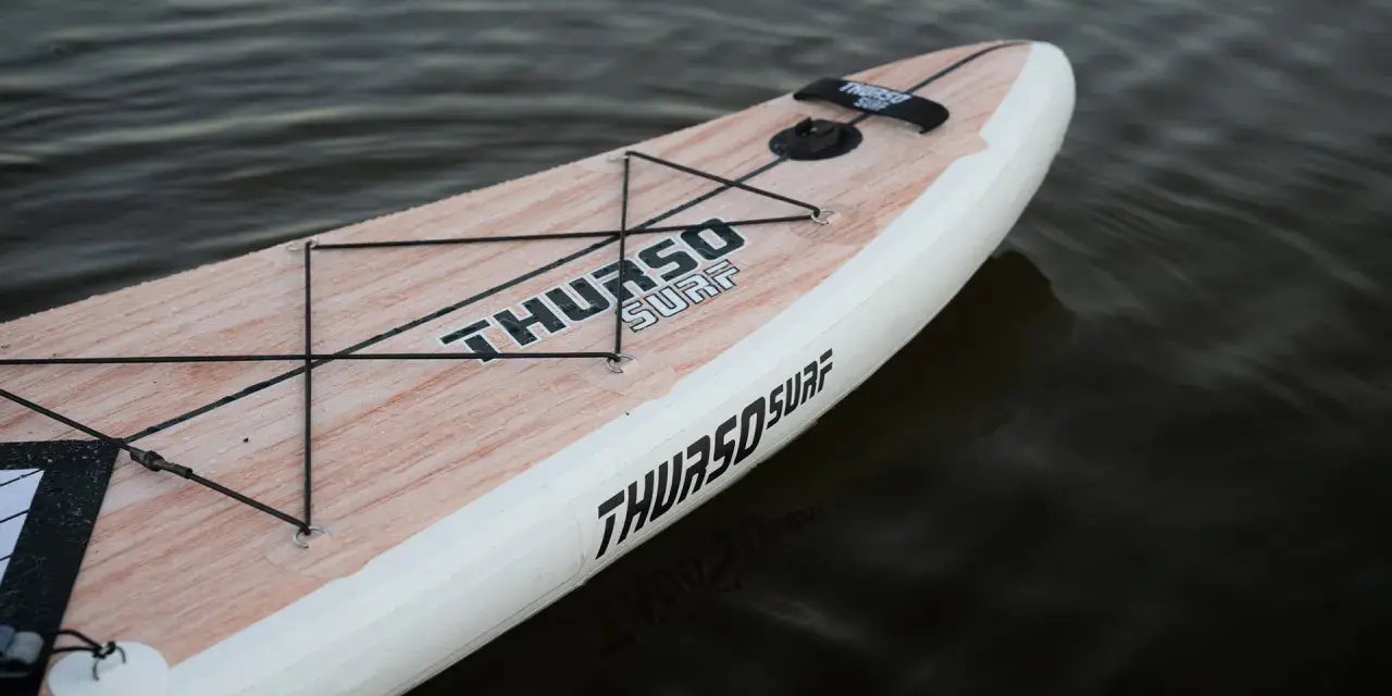Ein aufblasbares SUP liegt im Wasser. Das Board hat eine Holzoptik und das Logo "THURSO SURF" ist gut sichtbar. Perfekt zum SUP lagern.