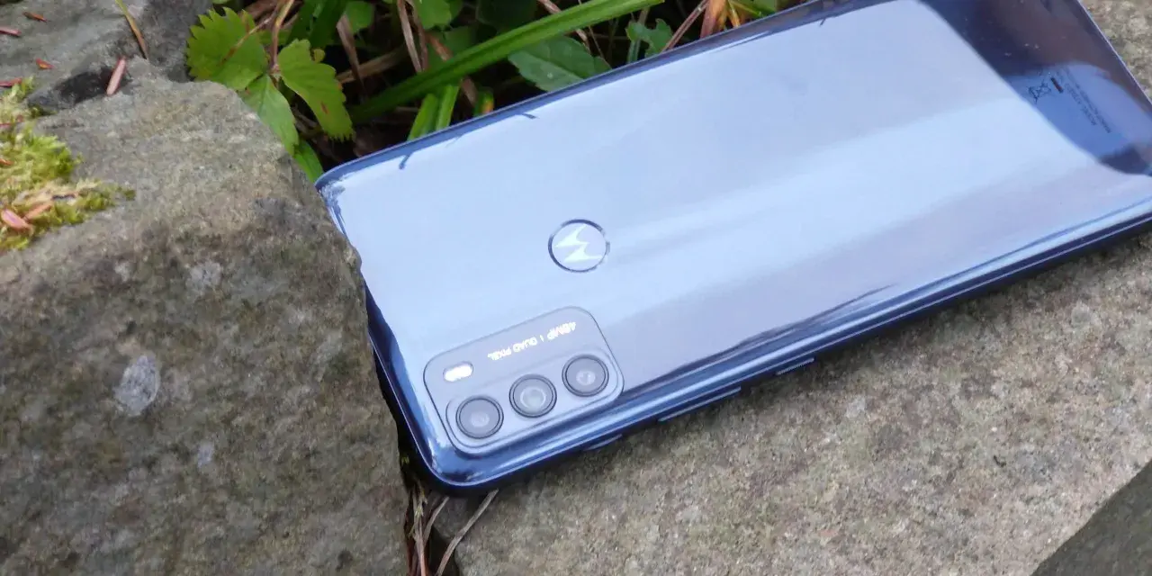 Błyszczący tył telefonu **Moto G 50** z potrójnym aparatem, leżący na kamieniach wśród zieleni.