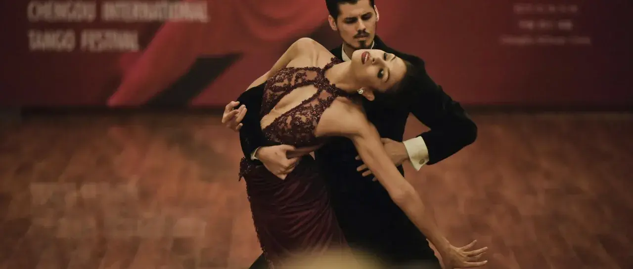 Para tańczy tango, jeden z rodzajów tańca towarzyskiego. Kobieta w bordowej sukni i mężczyzna w czarnym fraku wykonują skomplikowaną figurę.