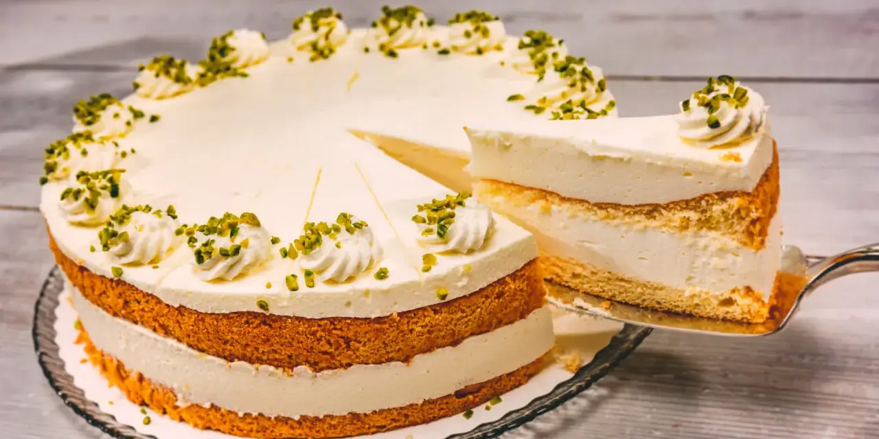 Eine Buttermilch Zitronen Torte mit cremiger Füllung und Pistazien-Deko. Ein Stück wird gerade herausgeschnitten.