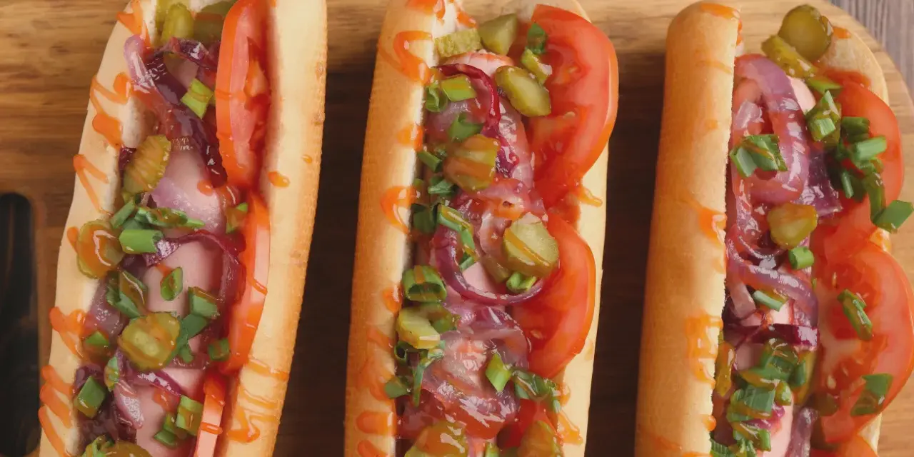 Trzy apetyczne hot dogi z frytkownicy powietrznej, z pomidorami, ogórkami, cebulą i szczypiorkiem, polane sosem.
