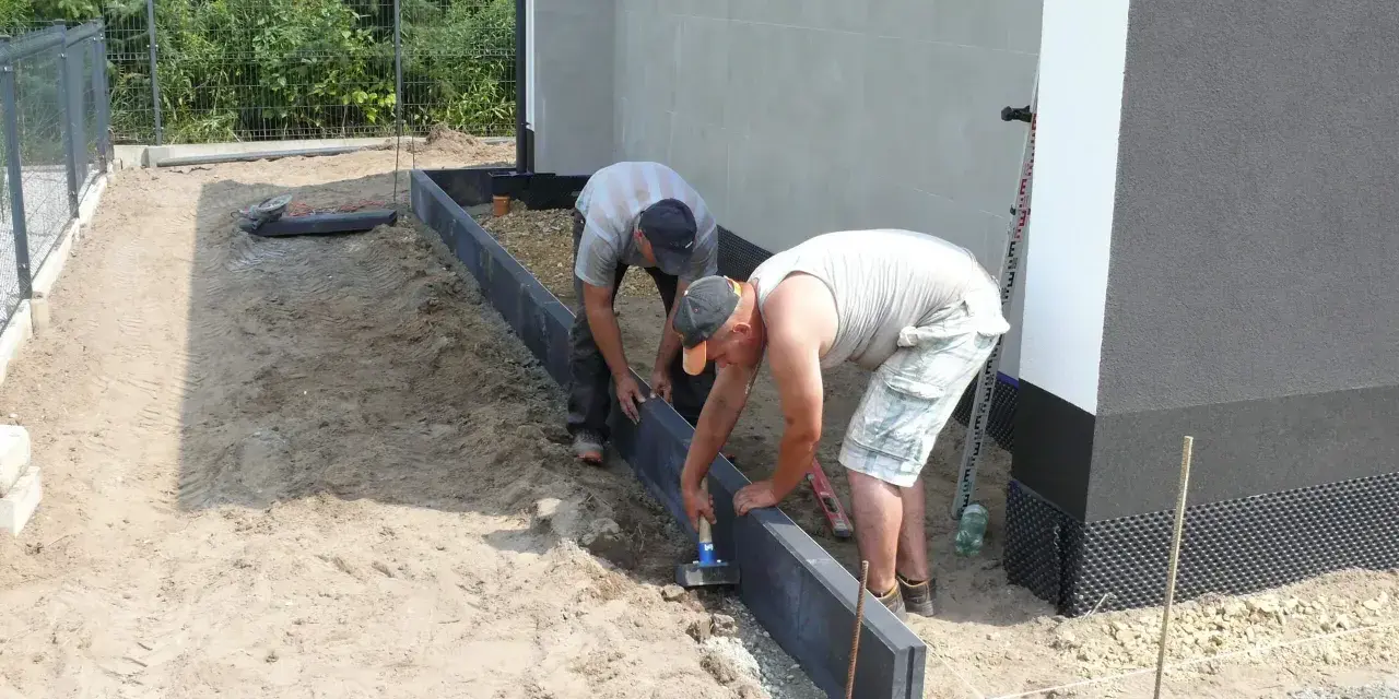 Dwóch mężczyzn układa obrzeża pod kostkę. Suchy beton pod kostkę jest przygotowywany do dalszych prac.