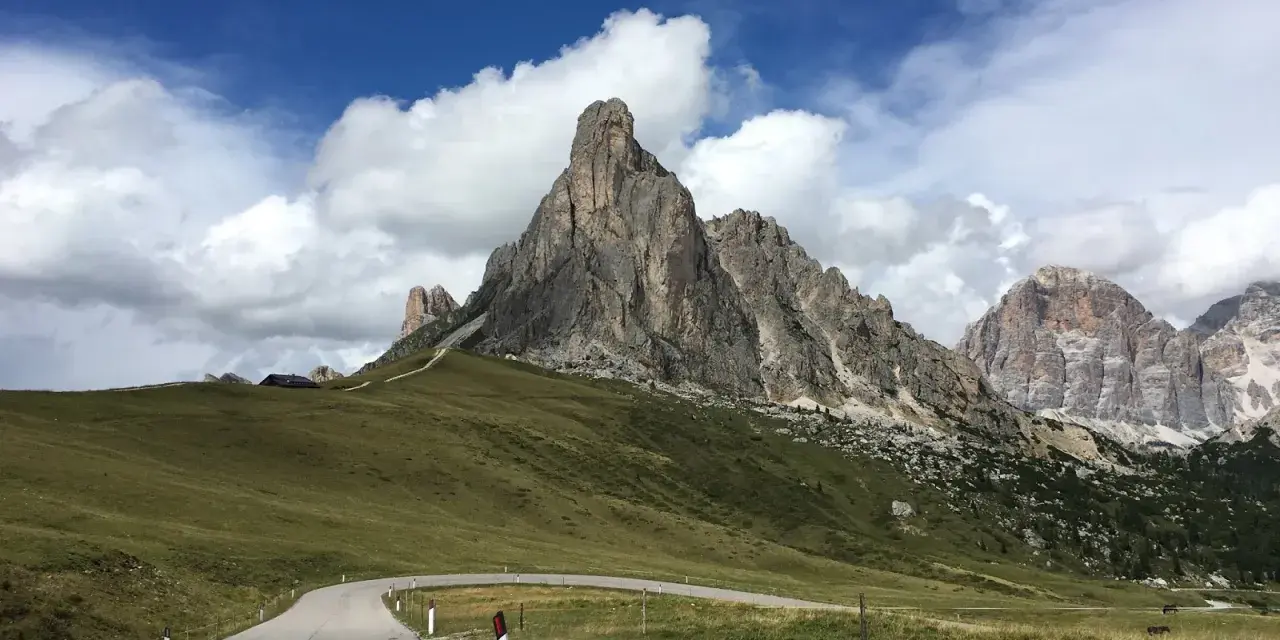 Strada tortuosa verso le Dolomiti, perfetta per una gita in moto. Montagne imponenti e cielo azzurro con nuvole bianche.