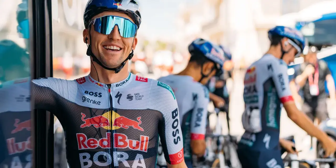 Cyclist smiles on a vuelta rest day, wearing a BORA-hansgrohe kit with Red Bull logos. Other team members and bikes are visible.
