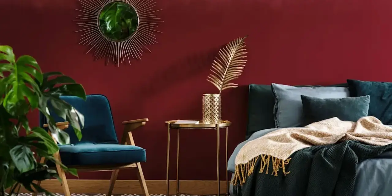 Habitación con pared de pintura a la tiza roja, sillón azul, cama con mantas y cojines, espejo sol y planta.