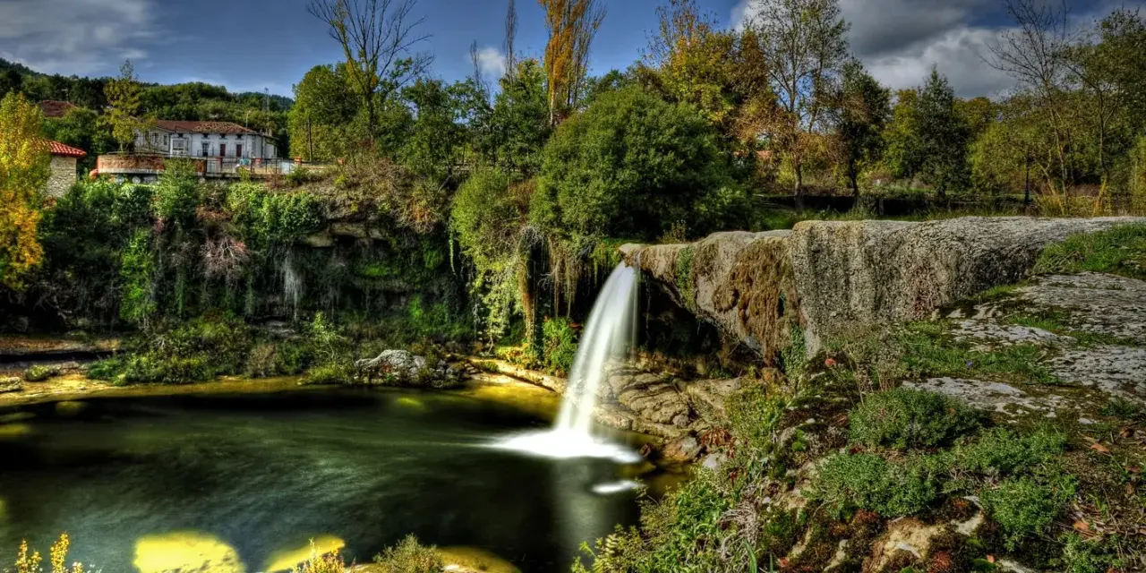 Cascadas en España: una casa rural se asoma entre la vegetación exuberante junto a una cascada que cae en un estanque de aguas verdes.