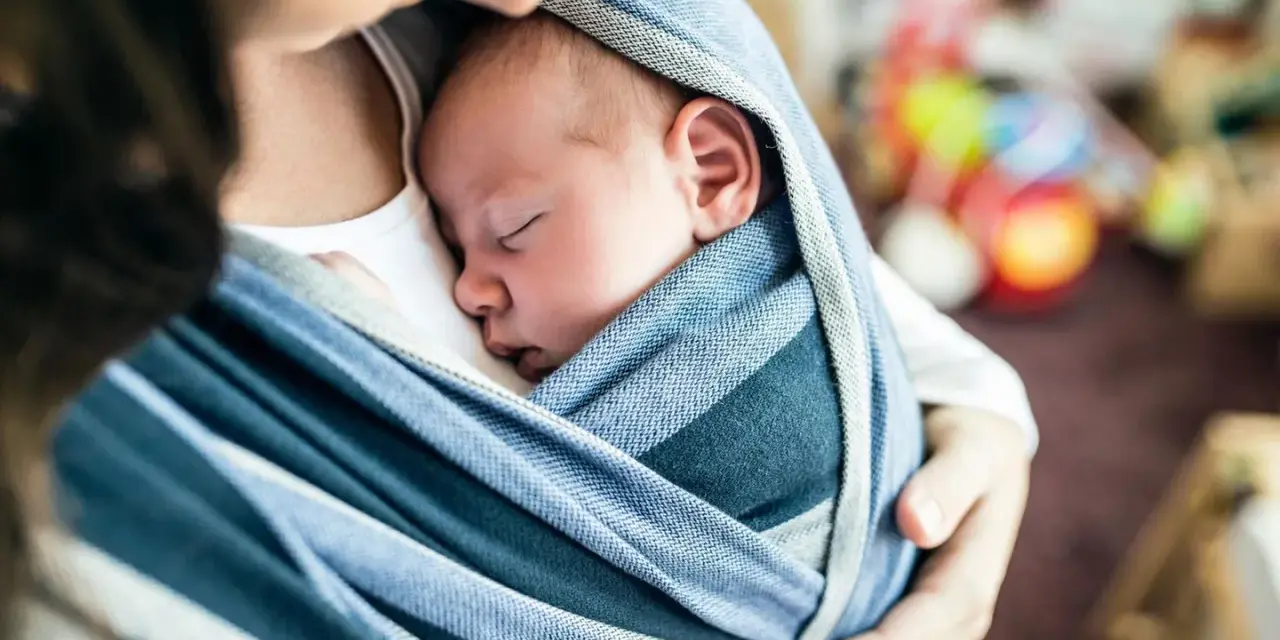 Ein Baby schläft friedlich in einem blauen Tragetuch. Der sanfte Tagesrhythmus des Babys wird durch die Nähe zur Mutter unterstützt.
