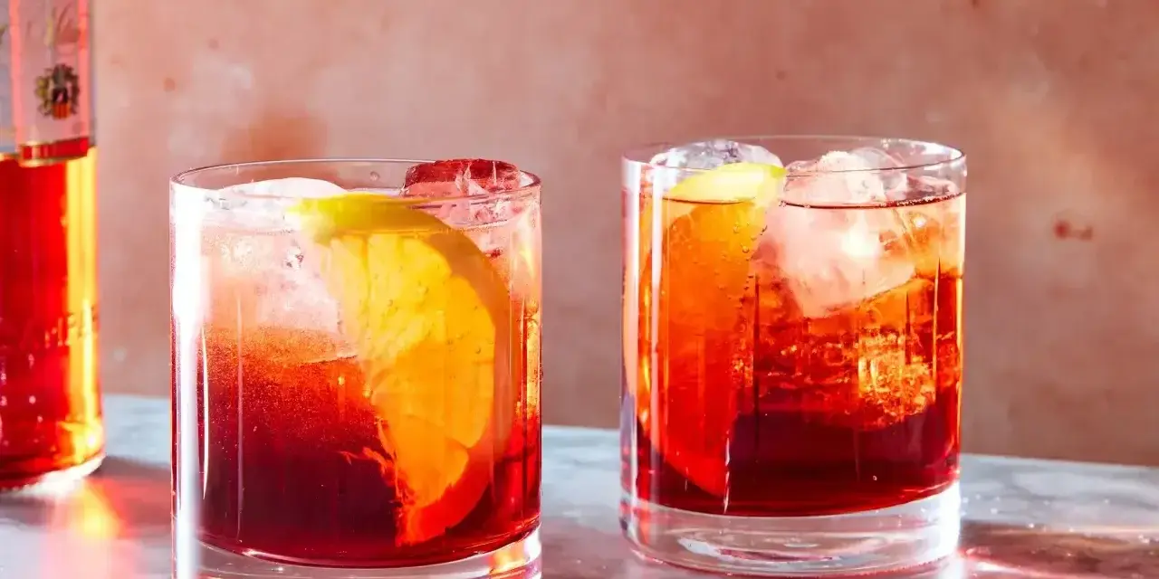 Dwa drinki Negroni z pomarańczą i lodem, obok butelki Campari. Idealny drink negroni przepis na lato.
