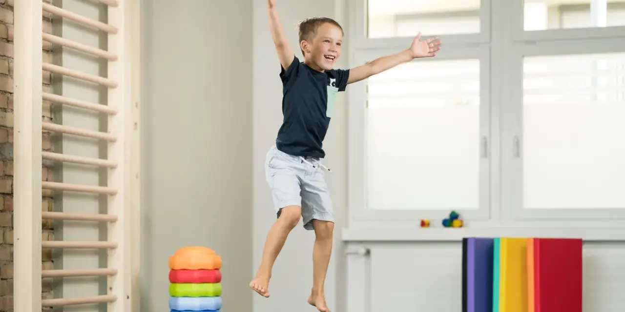 Fröhlicher Junge springt auf Trampolin, Teil der kinder physiotherapie. Kletterwand und bunte Matten im Hintergrund.