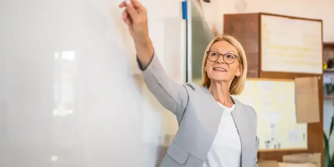 Nauczycielka z uśmiechem wskazuje markerem na tablicę, przygotowując wniosek o dokonanie oceny pracy nauczyciela.