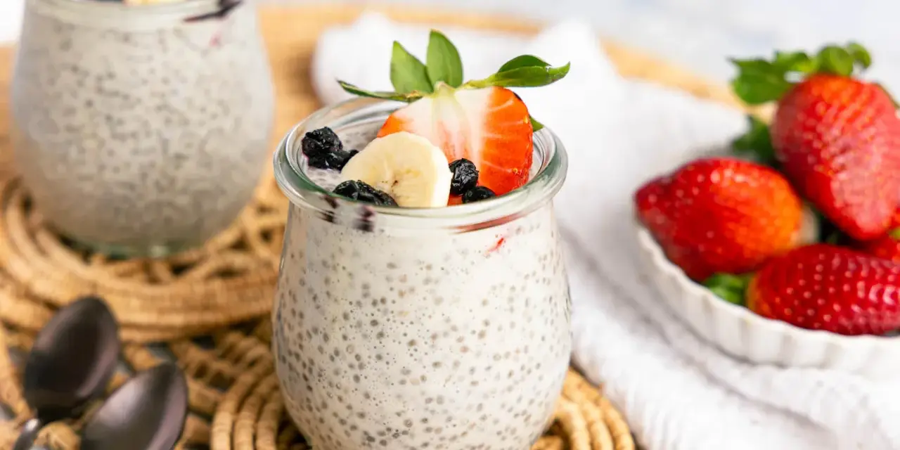 Zwei Gläser mit cremigem Chia Pudding, garniert mit Erdbeeren, Bananen und Blaubeeren. Daneben eine Schale frischer Erdbeeren.