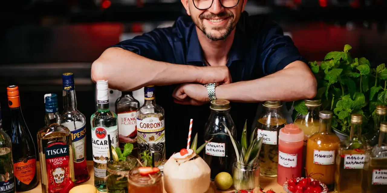 Uśmiechnięty mężczyzna w czapce i okularach za ladą pełną alkoholi i owoców. To przyszłość: drink bar zamiast barmana.