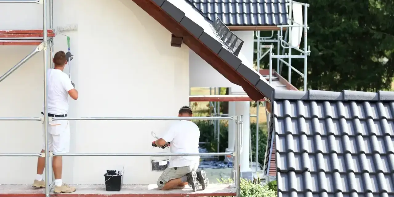 Dwóch mężczyzn na rusztowaniu maluje białą elewację domu. Sprawdź, jaka jest malowanie elewacji cena.